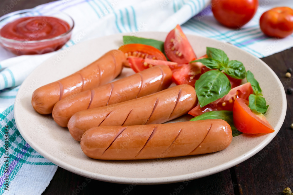 Juicy sausages with tomatoes and basil leaves on a plate on a dark wooden background. Top view. Juicy sausages with tomatoes and basil leaves on a plate on a dark wooden background. Top view.