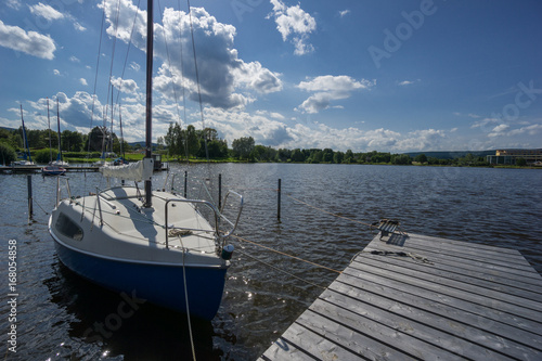 Ein kleines Segelschiff am Steg auf dem See und Himmel mit Wolken, wunderschön, wie das Wasser glitzert 