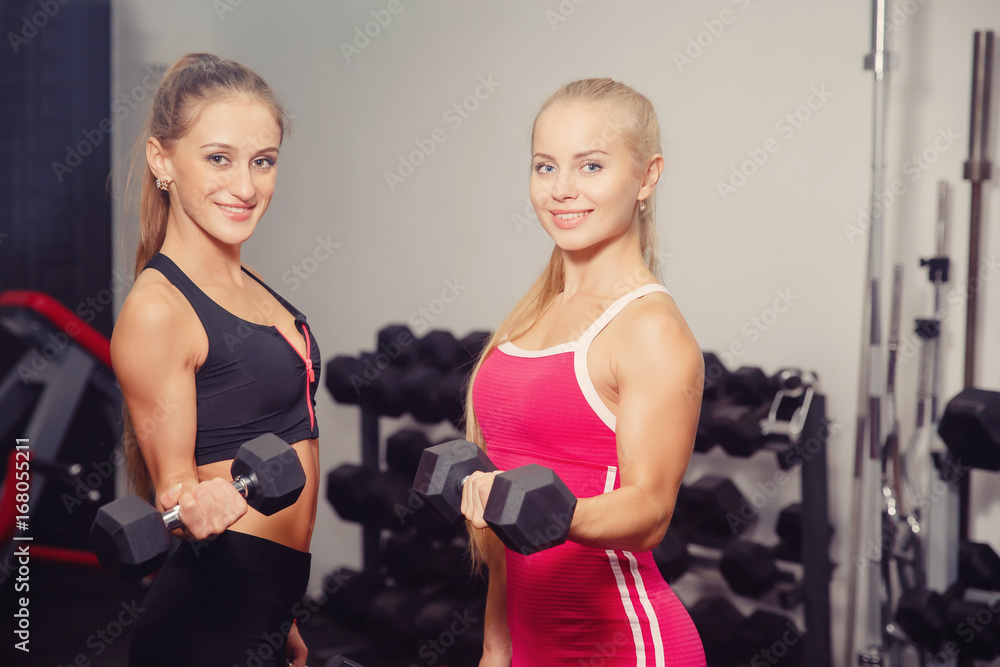 Close-up two girls smiling holding dumbbells in their hands and performing an exercise on the biceps, bending the arms at the elbow. Concept group sports with a friend. Close-up two girls smiling holding dumbbells in their hands and performing an exercise on the biceps, bending the arms at the elbow. Concept group sports with a friend.