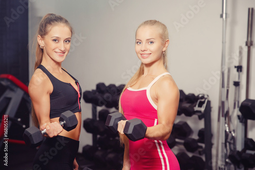 Close-up two girls smiling holding dumbbells in their hands and performing an exercise on the biceps, bending the arms at the elbow. Concept group sports with a friend.