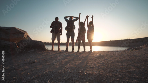 Foto Back view of four people with backpacks posing with hands up on coastline in back lit
