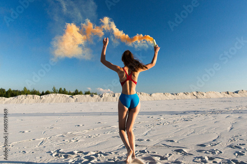 Girl in blue swim-suit dances with orange smoke on white beach