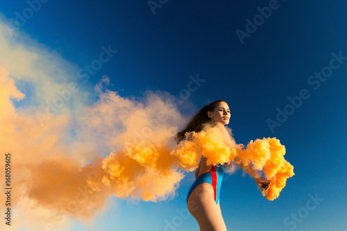 Girl in blue swim-suit dances with orange smoke on white beach