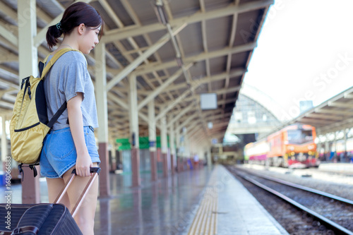 Young girl asian backpack traveler waiting with carrying hold suitcase luggage booking ticket in train station railway platform vacation time in holiday relaxation.