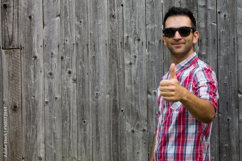 Portrait of man giving thumbs up sign, wearing sunglasses, smiling while standing against wooden background. photo