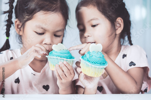 Quadro su tela Two cute asian child girls having fun to eat blue cupcake together in vintage co