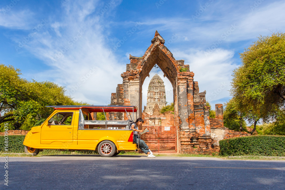 Obraz premium Young asian female traveler with backpack traveling sitting on taxi or Tuk Tuk and see map travel with old temple (Wat Mahathat) background, Ayutthaya Province, Thailand