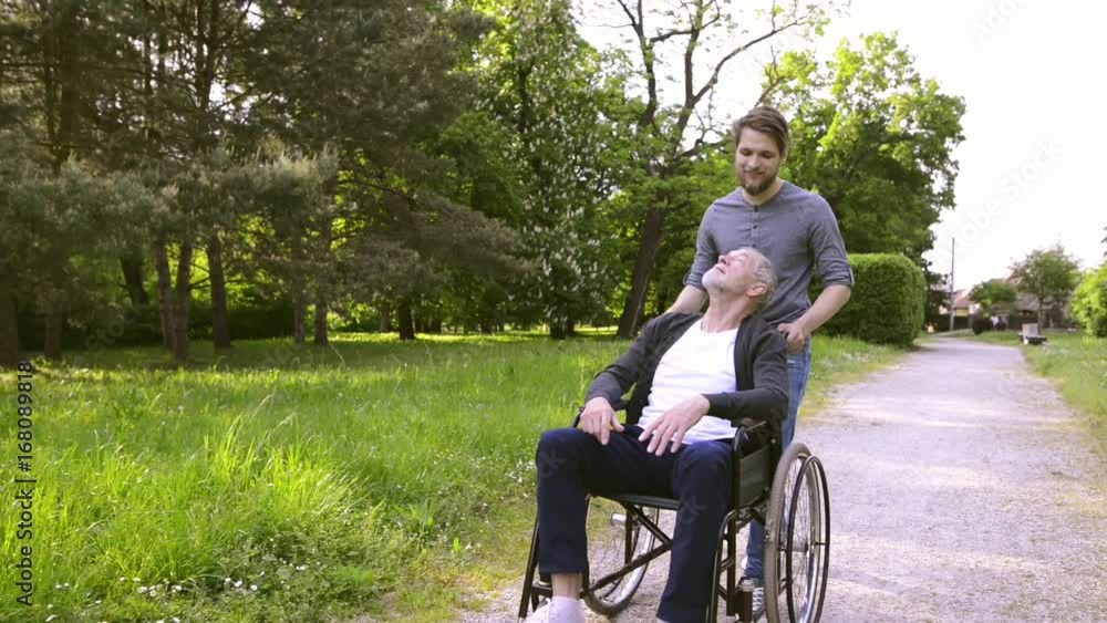 Hipster son walking with disabled father in wheelchair at park. Hipster son walking with disabled father in wheelchair at park.