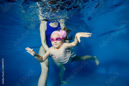 Little girl swims underwater in the pool on a blue background, and mom helps her. Portrait. Close-up. The view from under the water. Landscape orientation