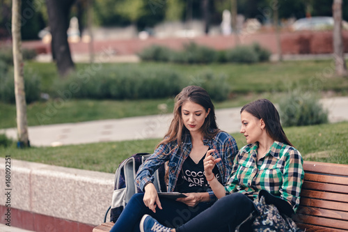 Modern girls sitting on a wooden bench. Talking seriously about their school supplies. Worried about the school supplies list. Looking at her sneakers. School backpacks are on bench. Back to school