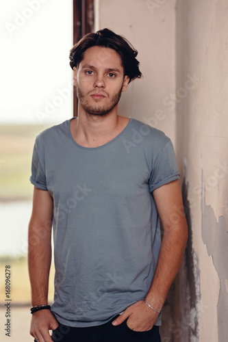 Trendy young man posing in grey t-shirt and black jeans posing like a model. Standing near the window in the evening sunlight. Listening to someone’s question and looking uncertain. © Damian
