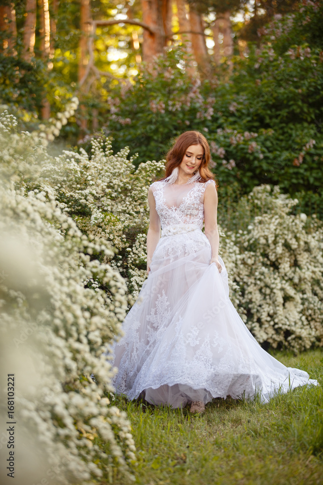 Beautiful redhead Bride in blooming garden playing with her dress. Portrait outdoor in sunset light. Pretty young caucasian redhead girl