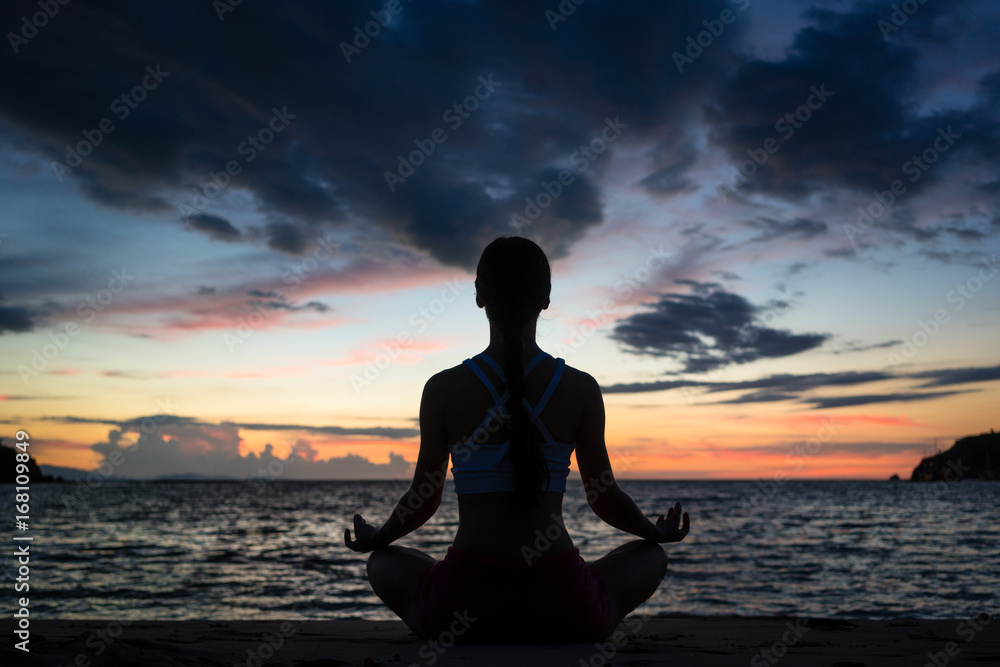 Full length rear view of a fit woman sitting in lotus position while practicing yoga on the beach at twilight during summer vacation in Flores Island, Indonesia