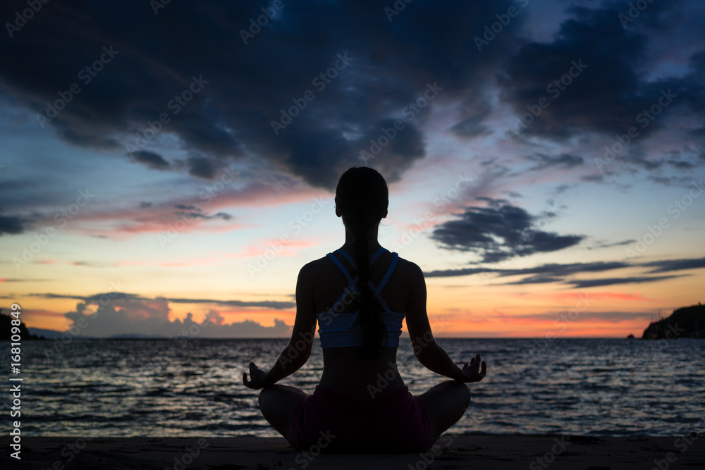Full length rear view of a fit woman sitting in lotus position while practicing yoga on the beach at twilight during summer vacation in Flores Island, Indonesia Full length rear view of a fit woman sitting in lotus position while practicing yoga on the beach at twilight during summer vacation in Flores Island, Indonesia