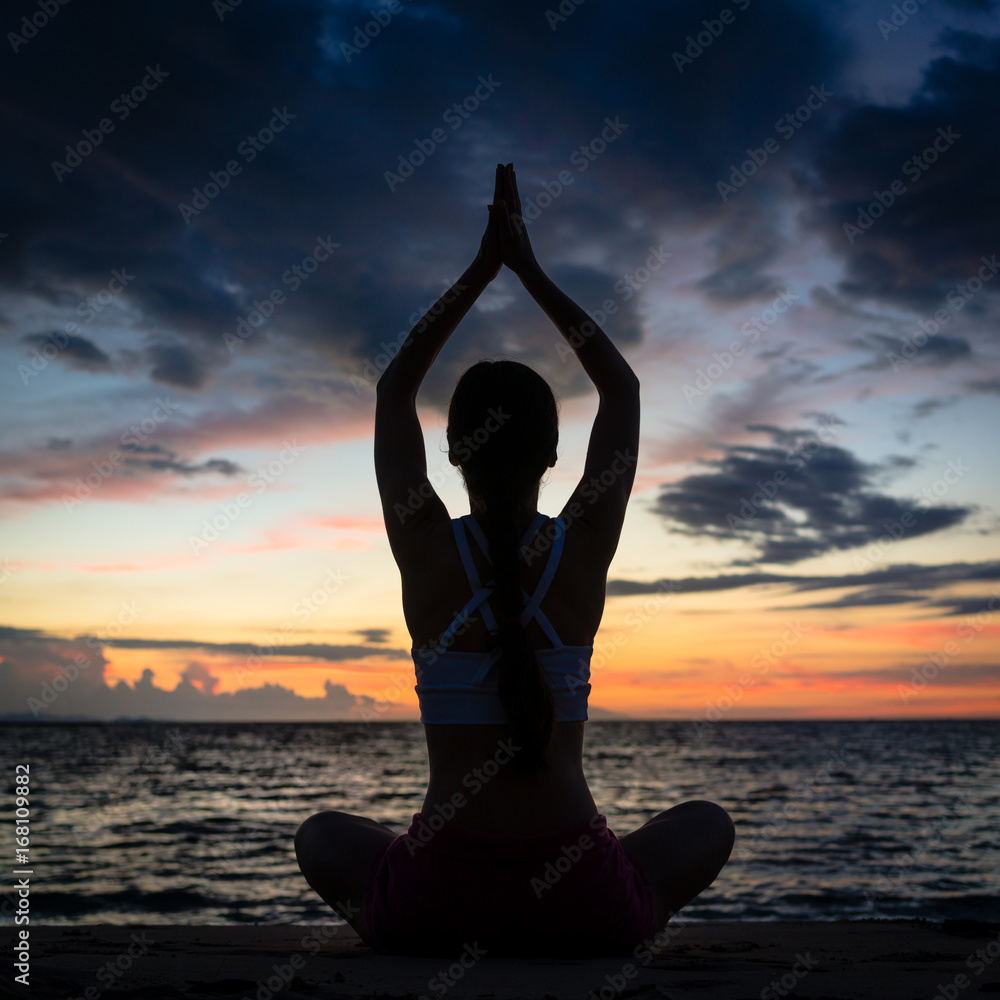 Full length rear view of a fit woman sitting in lotus position while practicing yoga on the beach at twilight during summer vacation in Flores Island, Indonesia