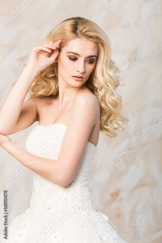 fashionable gown, beautiful blonde model, bride hairstyle and makeup concept - portrait of romantic lady in white festive wedding dress, slender pretty woman stand indoors on light background