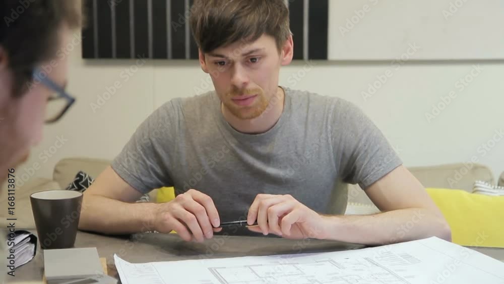 Collaborative work images design drawings on meeting, discussing inside office. Two Managers workers discuss business project, look on sheets papers with drawings. man with beard in light shirt sits