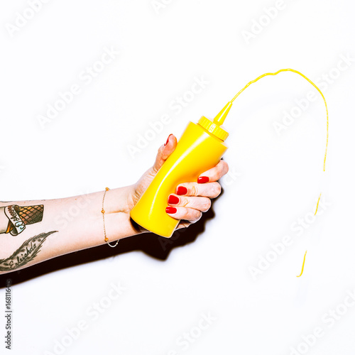 tattooed arm squeezing yellow mustard bottle with mustard squirt, on white background, studio