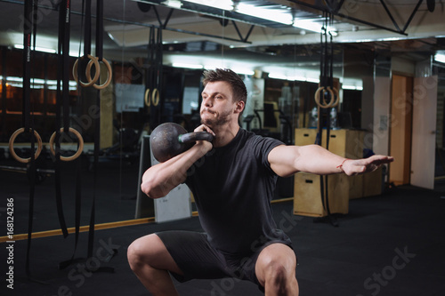 Pretty young man doing squats with dumbbell at the gym