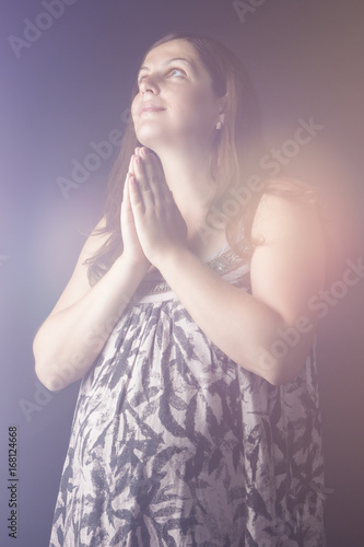 Portrait of Praying Pregnant Caucasian Female. Posing With Hands on Belly Against Black Background.
