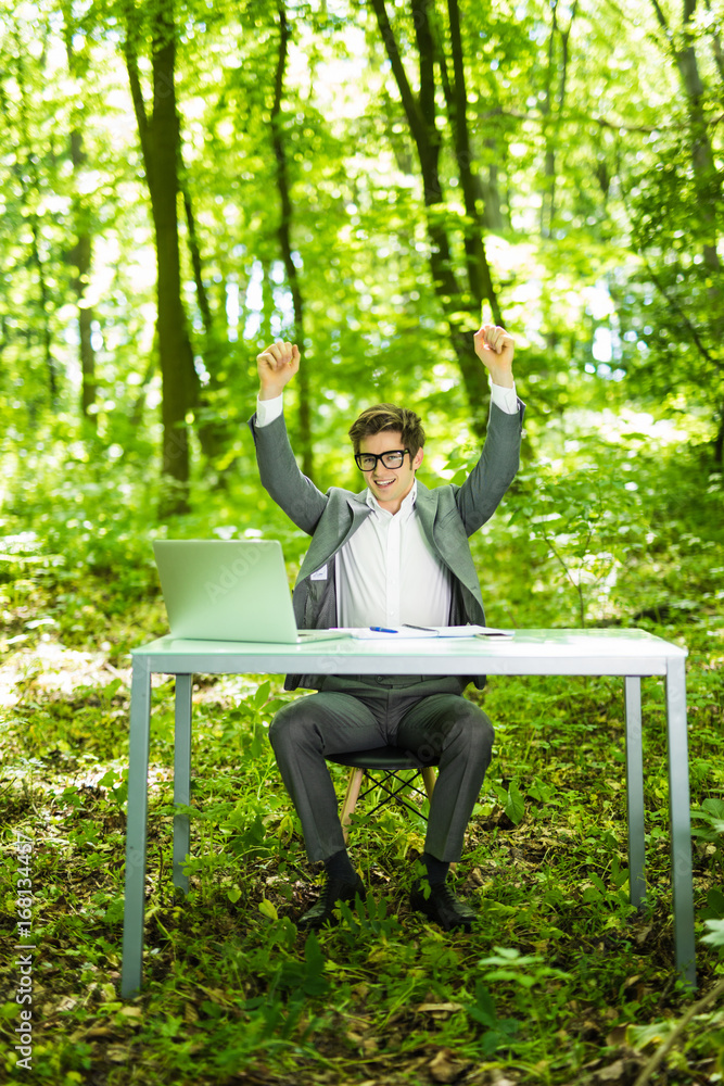 Portrait of young handsome succesful business man in suit working at laptop at office table with raised hands of victory in green forest park. Business concept. Portrait of young handsome succesful business man in suit working at laptop at office table with raised hands of victory in green forest park. Business concept.