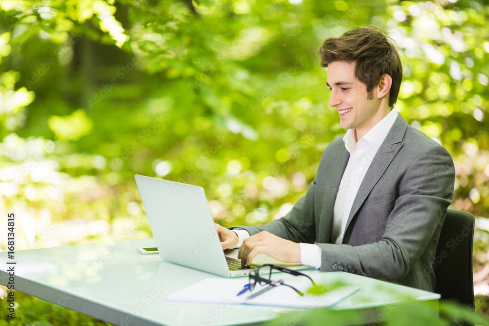 Side Portrait of young handsome business man in suit working at laptop at office table in green forest park. Business concept. Side Portrait of young handsome business man in suit working at laptop at office table in green forest park. Business concept.