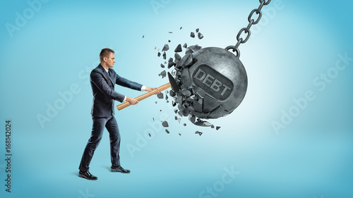 A small businessman smashes a giant swinging iron ball with a word DEBT on it using a hammer. photo