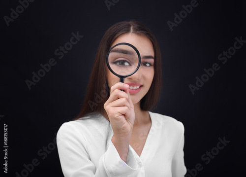 Beautiful young woman with magnifier on dark background