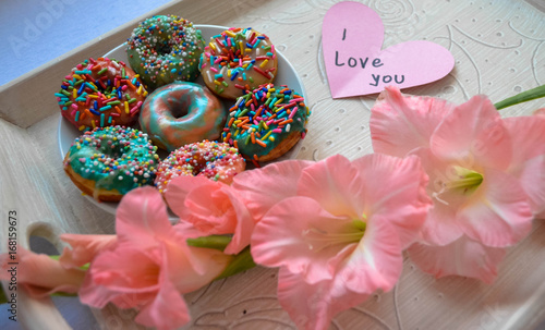 The husband surprises his wife for the anniversary with flower and donuts. Surprise has a note ''I love you'' next to gladiola flower.
