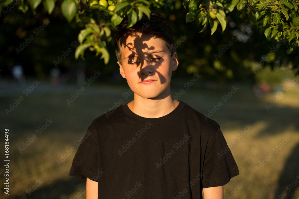 Portrait of young dreamy teen boy with eyes closed in garden. Beautiful shadow from the leaves on the face