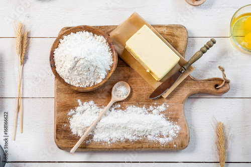 Baking ingredients in rural kitchen - dough recipe ingredients (eggs, flour, milk, butter, sugar) and rolling pin on wooden white table. Top View