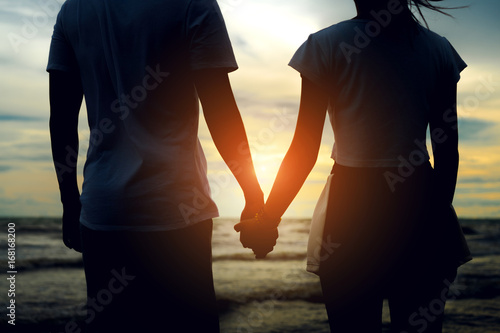 Young asian man holding hand with girl romantic together relationship valantine lover day at beach vacation time leisure back view sunset evening.