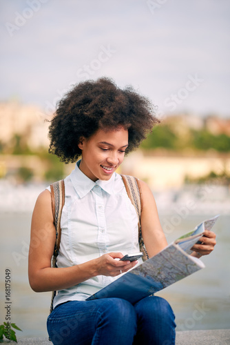 Mixed race female tourist looking at map and using smart phone while sitting by the river