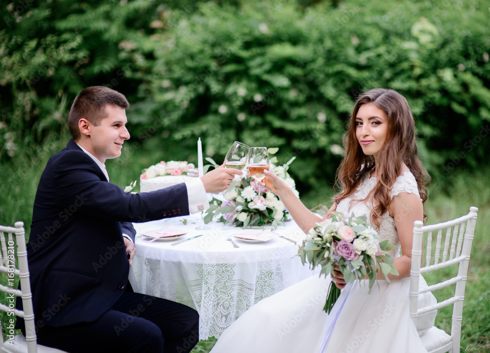 Wedding couple drinks champagne sitting at the table in a green garden Wedding couple drinks champagne sitting at the table in a green garden