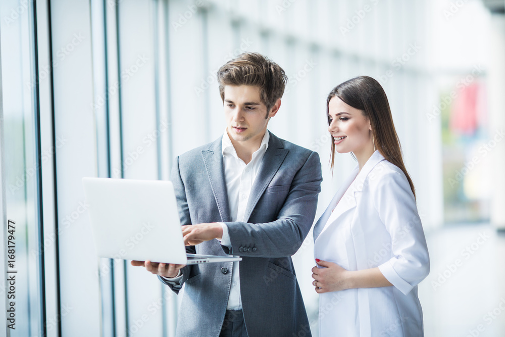Business man and business woman discussing new project on the laptop atanding against in office room with panoramic windows Business man and business woman discussing new project on the laptop atanding against in office room with panoramic windows