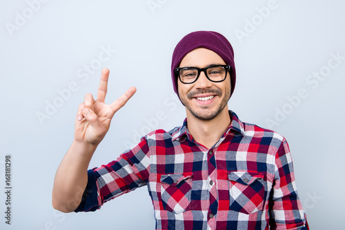 Peace! Smiling young nerdy stylish student hipster is gesturing vsign on pure background in black trendy glasses and hat, casual bright checkered shirt photo
