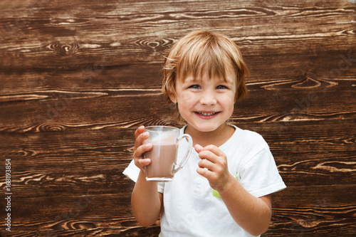 the kid drinking cocoa at home against the background of a wooden wall.