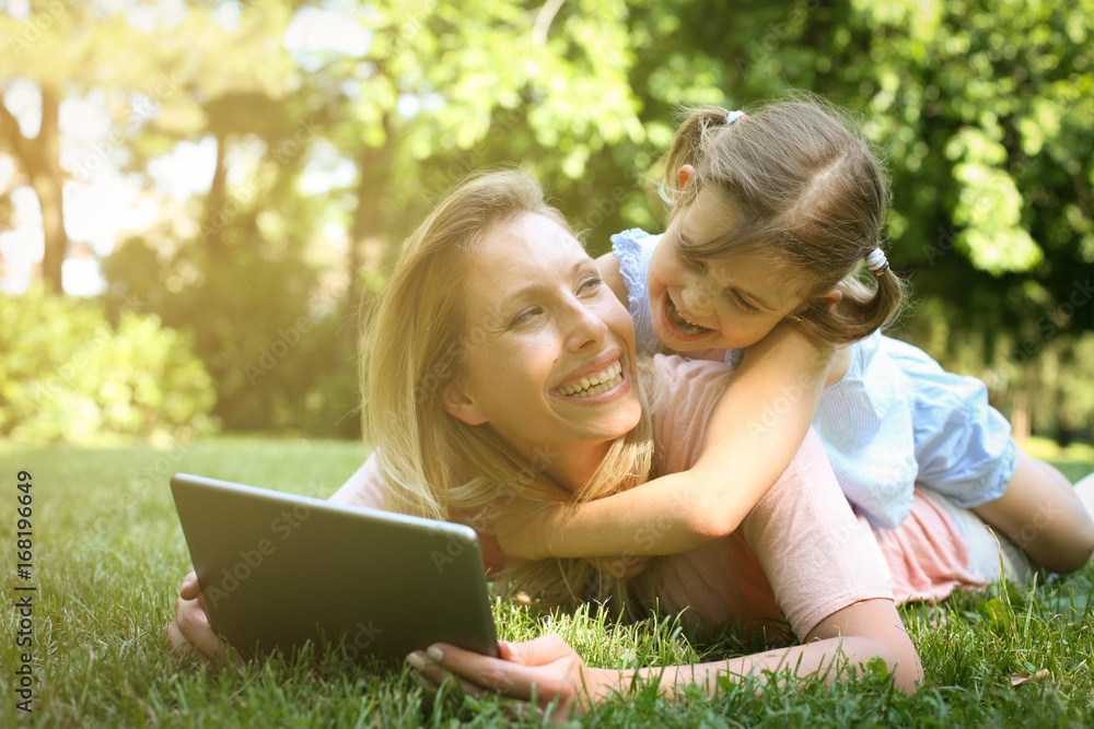Mother and daughter outdoors in a meadow. Mother with her daughter lying on grass and using digital tablet together. Mother and daughter outdoors in a meadow. Mother with her daughter lying on grass and using digital tablet together.