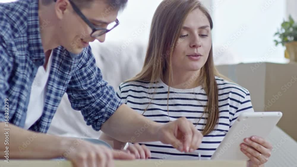 Medium shot of happy young woman with tablet giving instructions to cheerful boyfriend screwing bolts into wooden furniture Medium shot of happy young woman with tablet giving instructions to cheerful boyfriend screwing bolts into wooden furniture