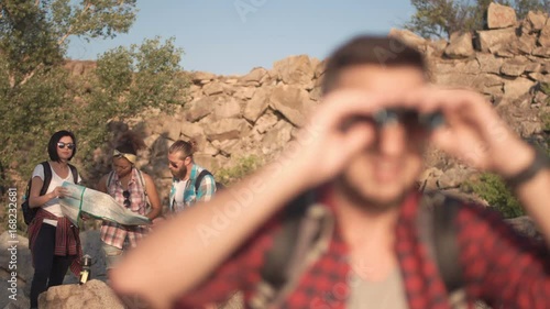 Young man looking at friends with map behind him and then looking in binocular while traveling.
