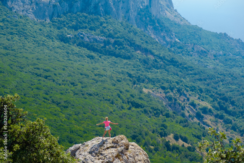 A young girl stands on a rock with arms outstretched in the background of a mountain landscape.