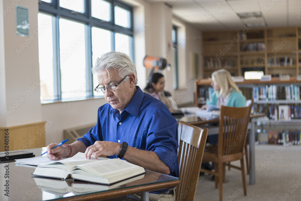 Older man reading book in library and writing notes Older man reading book in library and writing notes