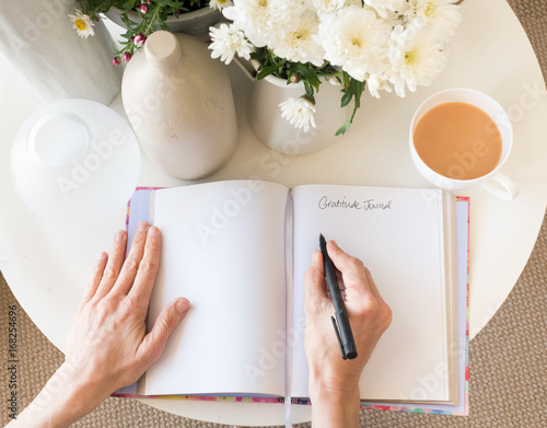 High angle cropped view of woman's hands writing in gratitude journal at desk with tea and flowers (selective focus) photo