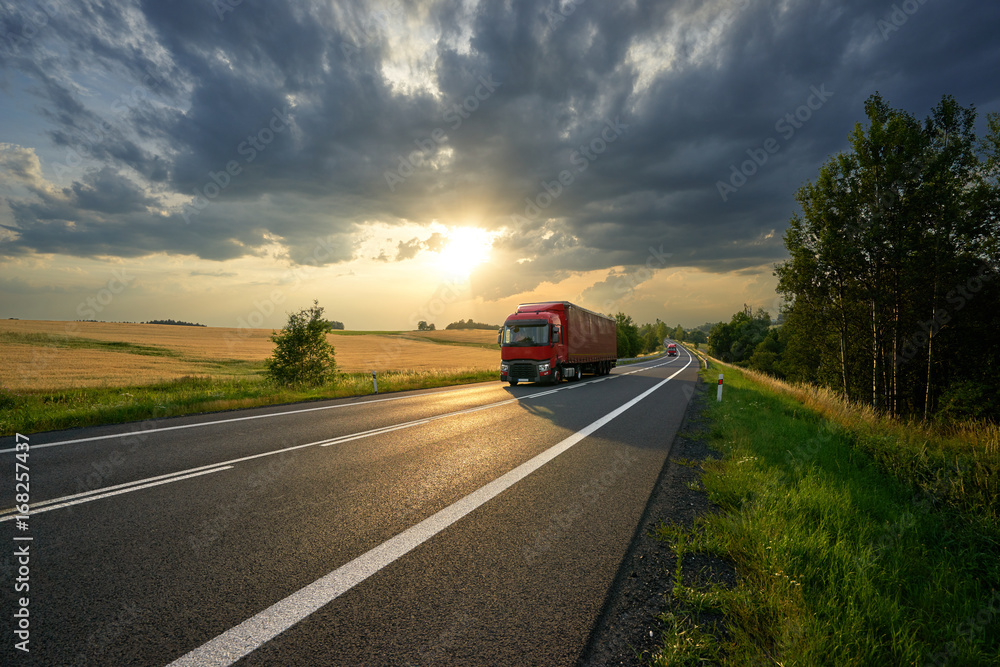Red trucks arriving on the asphalt road along a field with golden grain in rural landscape at sunset with dark clouds