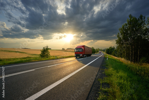 Obraz na płótnie Red trucks arriving on the asphalt road along a field with golden grain in rural landscape at sunset with dark clouds w salonie