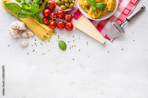 Italian food cuisine and ingredients on white concrete table. Spaghetti Tagliatelle olives olive oil tomatoes parmesan cheese garlic pepper and basil leaves and checkered tablecloth. 
