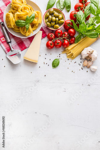 Italian food cuisine and ingredients on white concrete table. Spaghetti Tagliatelle olives olive oil tomatoes parmesan cheese garlic pepper and basil leaves and checkered tablecloth.