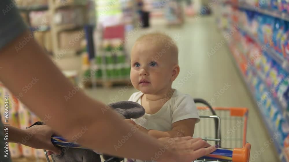 Little baby sitting in a grocery cart in a supermarket holding a toy, while her mother is pushing the cart walking among choosing products. Family shopping with a little child Little baby sitting in a grocery cart in a supermarket holding a toy, while her mother is pushing the cart walking among choosing products. Family shopping with a little child