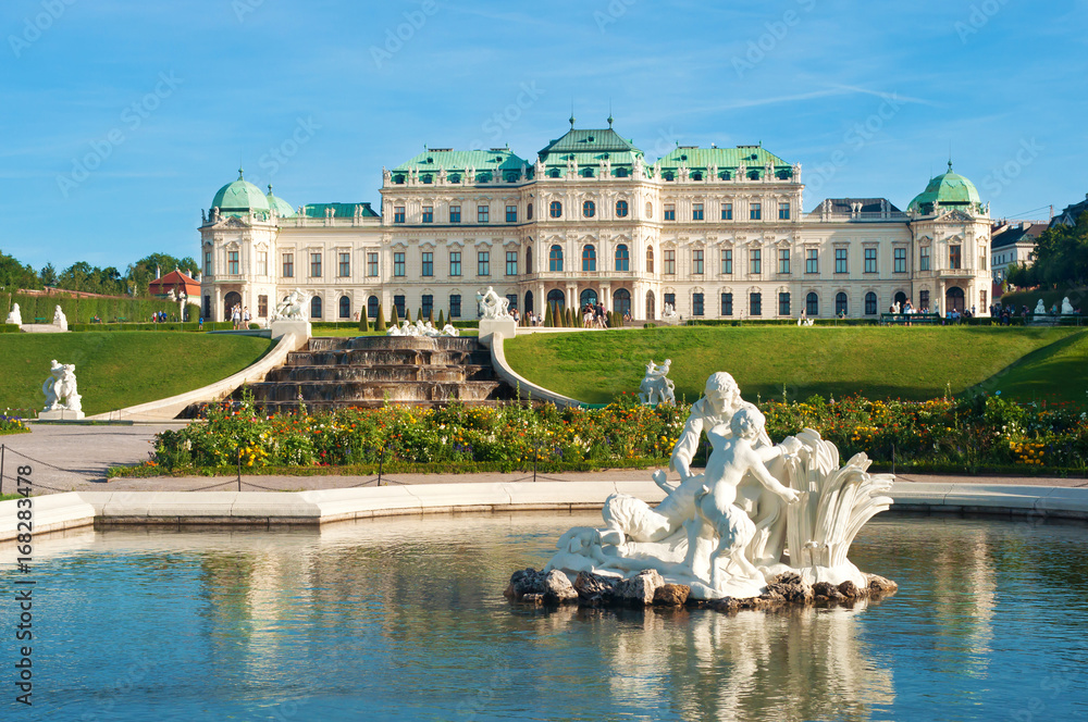 View on Belvedere palace, fountain, flowerbed with blooming flowers and several white statues of people in the water of a pond against a cloudless blue sky. Summer, Vienna