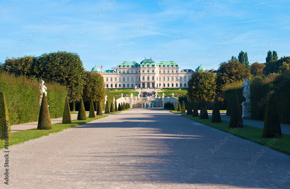 View on an empty solitary lane with trimmed conical bushes and green trees on both sides leading to Belvedere palace in the distance, against a cloudless blue vibrant sky. Summer, Vienna